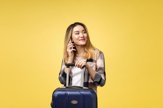 Portrait of happy asian female traveler with suitcase and looking at cellphone against isolated yellow background.