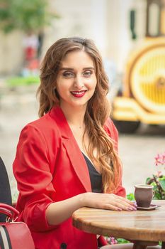 Portrait of pretty girl is sitting at cafe outside with cup of coffee at the table in the street.