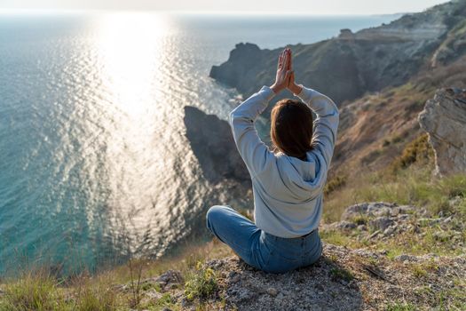 Woman tourist enjoying the sunset over the sea mountain landscape. Sits outdoors on a rock above the sea. She is wearing jeans and a blue hoodie. Healthy lifestyle, harmony and meditation.