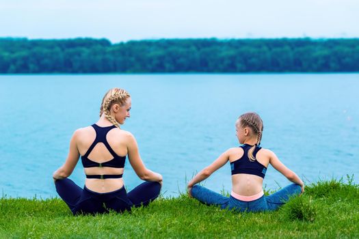 Mother and daughter doing yoga exercises on grass of the shore of the lake.