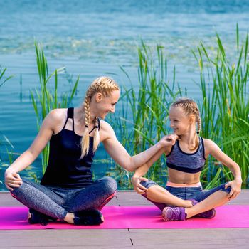 Portrait of mother and daughter sitting on yoga mat by the lake outdoors. Healthy and exercise concept.