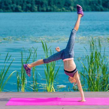 Fit sporty little girl in sportswear doing cartwheel exercise on the roll mat by the lake.