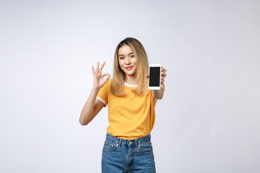 Young Asian woman wearing in yellow shirt is showing ok sign on white background, holding mobile phone, smiling