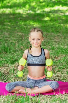 Little girl is training with apple dumbbells in the park. Healthy sport exercises.