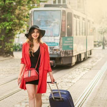 Beautiful young woman walking with a rolling suitcase on the tram track road in the old European city.