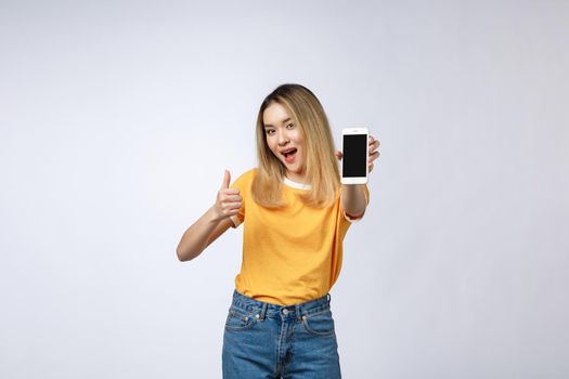 Young Asian woman wearing in yellow shirt is showing thumb up sign on white background, holding mobile phone, smiling