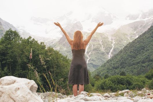 Back of caucasian reddish haired woman raising hands up in mountains in summer time feeling happiness and freedom. Bucket list