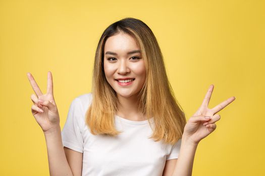 Pleased asian woman in t-shirt showing peace gestures and looking at the camera over yellow background.