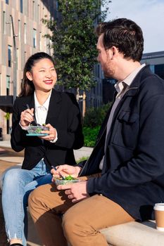 asian woman smiling and eating a salad with her caucasian partner in a park next to the office, concept of healthy fast food at work