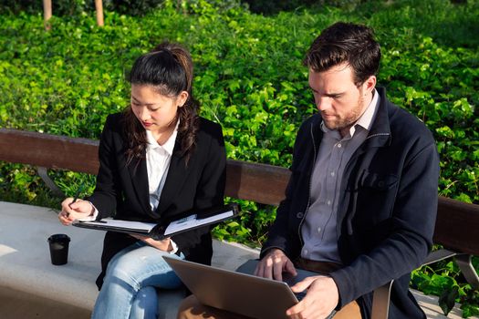 asiatic woman and caucasian man working outdoors with laptop in a park next to the office, concept of coworkers and diversity at work