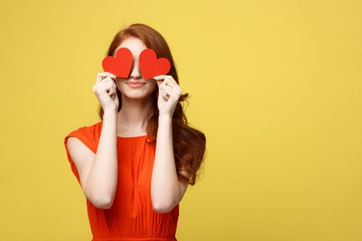 Lifestyle Concept: Attractive woman with beaming smile having two small red hearts in hands, closing eyes with paper heart symbols while standing over yellow background
