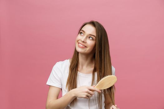 Smiling young woman combing hair and looking away isolated on pink.