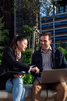 asian woman and caucasian man working outdoors with laptop in a park next to the office, concept of coworkers and diversity at work