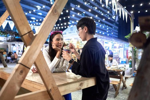 Non binary person feeding ice cream to her partner at urban night fair in Bangkok, Thailand