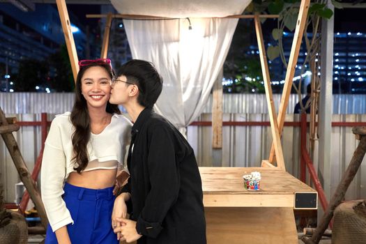 Non-binary person kissing her partner on the cheek in front of a booth at an evening fair