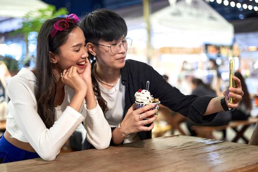 Lesbian couple eating an ice-cream terrine while taking a selfie at a night fair in Bangkok