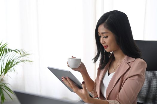 Young businesswoman drinking coffee and reading email on her digital tablet.