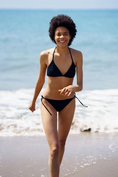 Happy black woman laughing while walking in bikini on the sand of the beach. Girl with afro hairstyle coming out of the water at the beach.