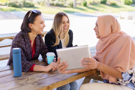 Group of positive young multiethnic women smiling and drinking coffee, to go while watching funny video on tablet, during break in outdoor cafe in park