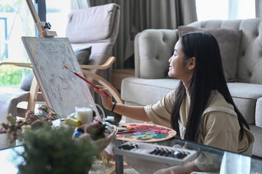 Cute little girl sitting on floor in living room and painting on canvas with watercolor.