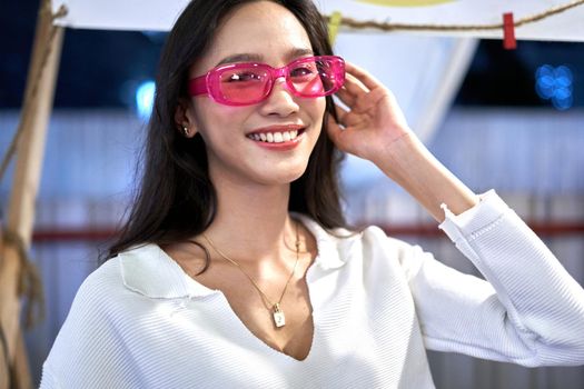 Portrait of an asian woman smiling sensually to the camera with pink glasses at an urban night fair