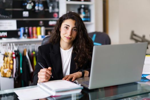 Young sports psychologist with laptop making notes and looking at camera during work in modern gym in daytime