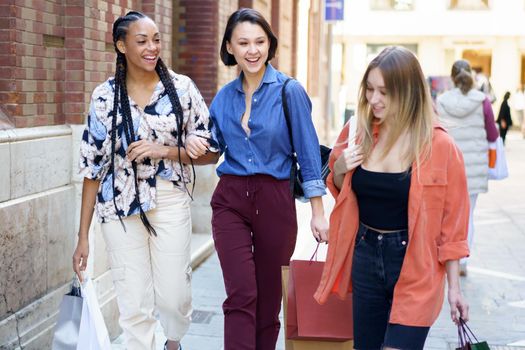 Group of optimistic multiethnic female friends with shopping bags strolling together on paved street near building after shopping in city