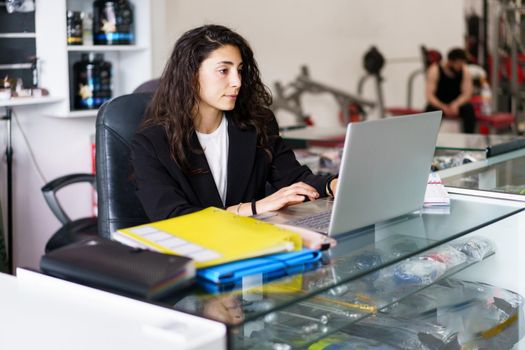 Young brunette in smart casual clothes browsing psychology article on netbook while sitting at desk with sports supplies during work in gym