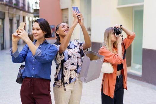 Content multiracial female friends taking pictures of city with modern smartphones and photo camera while standing on street near buildings