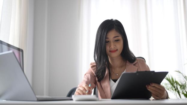 Smiling working woman holding clipboard and using calculator at office desk.
