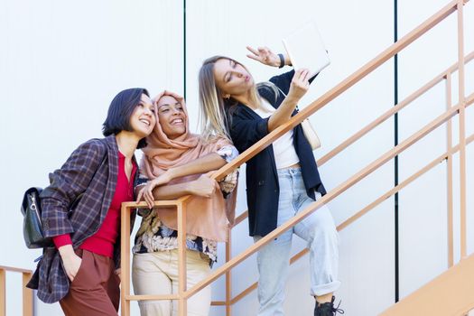 Low angle of delighted young multiracial female students in trendy outfit gesturing and smiling while taking selfie on tablet standing on stairs on city street