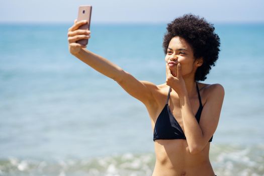 Black woman with afro hairstyle taking a selfie with a funny gesture on a tropical beach. Black girl enjoying her holiday on the beach.