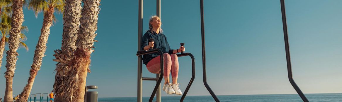 Adult female holding on uneven bars and raising her knees up on the sports field by the seashore