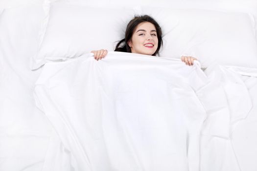 Top view photo of beautiful mixed-race girl lying on big white bed. Young pretty woman wearing pajamas. Girl lying on back, looking at camera and covering herself with blanket