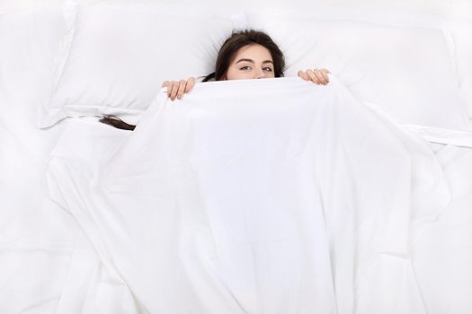 Top view photo of beautiful mixed-race girl lying on big white bed. Young pretty woman wearing pajamas. Girl lying on back, looking at camera and covering herself with blanket