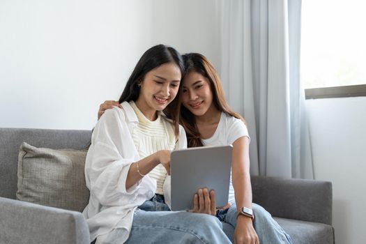 Happy young asian lesbian couple hugging having fun using digital tablet relaxing on couch at home. Two smiling women friends holding computer looking at screen enjoying surfing online watching videos.