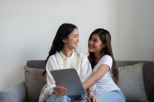 Happy young asian lesbian couple hugging having fun using digital tablet relaxing on couch at home. Two smiling women friends holding computer looking at screen enjoying surfing online watching videos.