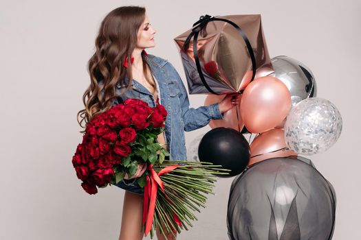 Pretty young slim brunette with wavy hair in denim jacket holding beautiful red roses bouquet and several colorful fashionable air balloons. She is smiling at her reflection in air balloon. Studio birthday party.
