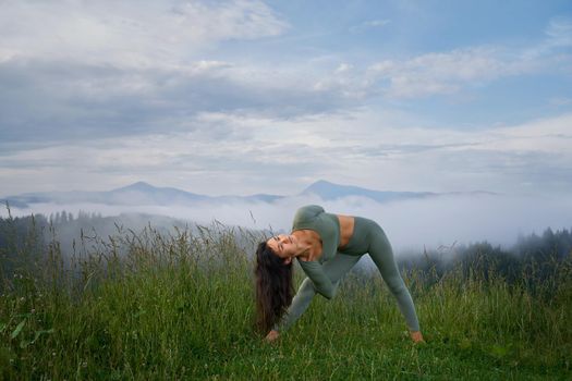 Young active woman with dark hair doing flexible exercises for body among summer mountains. Healthy and fit lady in sport clothes enjoying morning workout on fresh air.