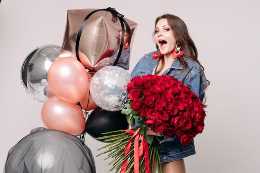Elegant young lady screaming while getting presents on holiday. Brunette girl in jeans jacket holding big bouquet of red roses tied with stripes. Surprised happy woman standing near colorful balloons.