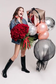 Full length studio shot of fashionable brunette in denim jacket and rubber boots holding bunch of red roses and air balloons. She is having birthday today. Isolate.