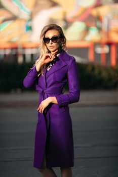 Young stylish woman in sunglasses posing at camera in sunny day. Blond girl in black blouse and violet coat holding her hands pockets. Elegant pretty lady in fashionable clothes standing outside.