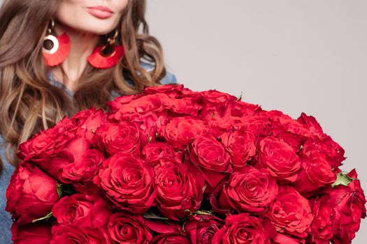 Close up of huge and elegant bouquet of red roses. Brunette woman with full lips and long hair holding flowers. Stylish lady in big gold and red earrings getting presents on her birthday.