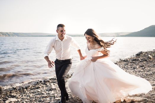 bride and groom in wedding dress happy and laughing run together against rays of setting sun in summer Bakota, Ukraine