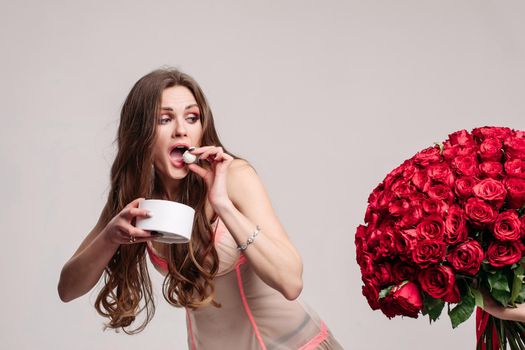 Funny woman in pyjamas eating sweets and running away from big beautiful bouquet of red roses. Pretty lady refusing proposal on grey isolated background. Concept of presents and gift.