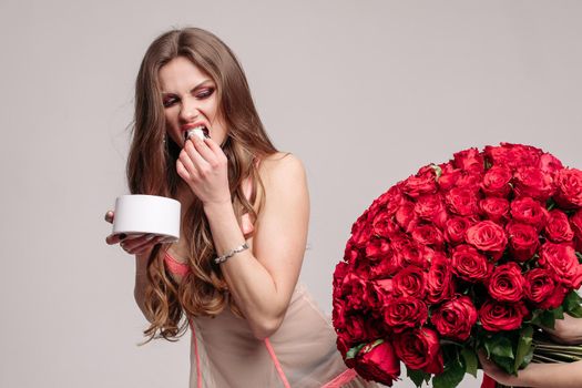 Funny woman in pyjamas eating sweets and running away from big beautiful bouquet of red roses. Pretty lady refusing proposal on grey isolated background. Concept of presents and gift.
