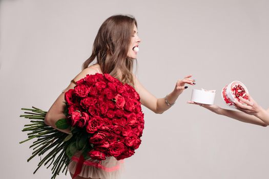 Studio shot of pretty brunette woman holding bouquet of gorgeous red roses. She is eating sweet and taking one more candy from the box in anonymous female hands. Isolate on white.