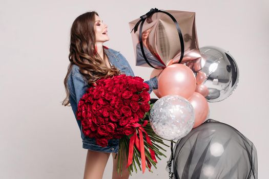 Pretty young slim brunette with wavy hair in denim jacket holding beautiful red roses bouquet and several colorful fashionable air balloons. She is smiling at her reflection in air balloon. Studio birthday party.