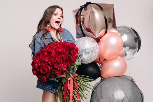 Elegant young lady screaming while getting presents on holiday. Brunette girl in jeans jacket holding big bouquet of red roses tied with stripes. Surprised happy woman standing near colorful balloons.