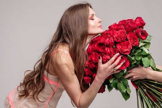 Studio portrait of stunning young brunette woman with make up wearing sexy night dress smelling a big bunch of red roses with closed eyes. Enjoying the smell of roses.